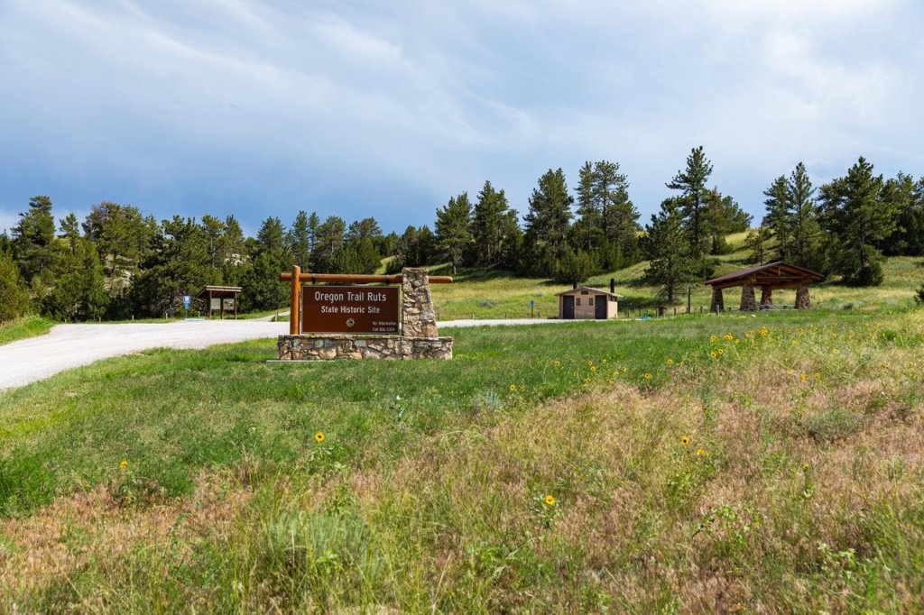 8 Historic Landmarks & Sites In Wyoming 3 A sign that reads Oregon Trail Ruts State Historic Site amidst a field of tall grass and wildflowers, and in the background, a rest area, interpretive sign and a forest of trees.