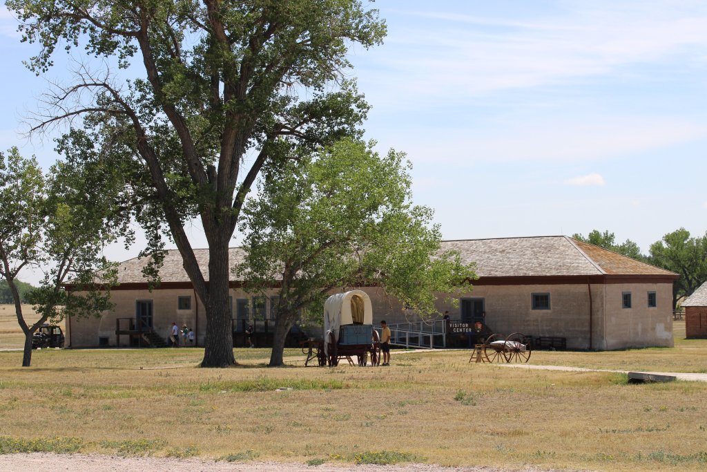 Historic building at Fort Laramie with large cottonwood trees, a covered wagon, and the visitor center in the background.