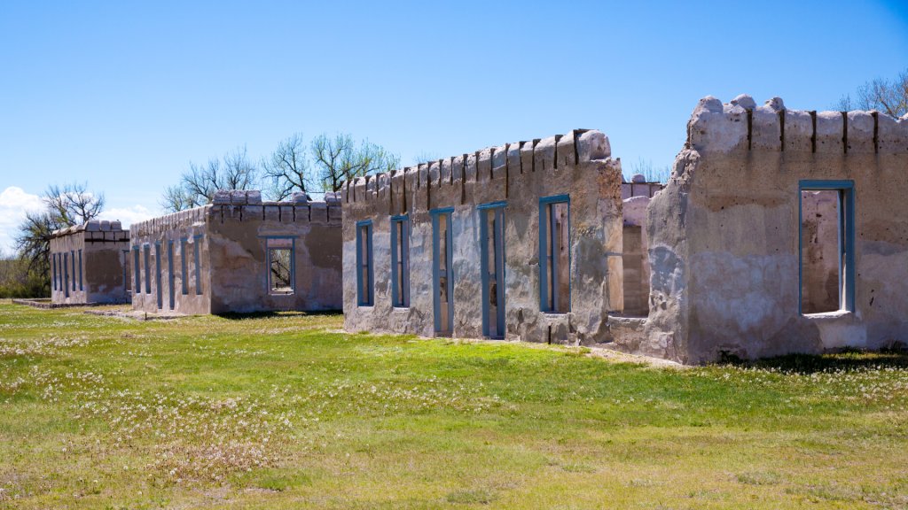 Crumbling adobe and stone ruins of historic Fort Laramie under a clear blue sky with scattered trees and green grass.