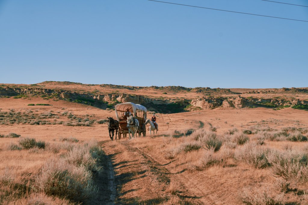 A man rides a horse-drawn covered wagon while a woman rides her own horse beside him across the West.