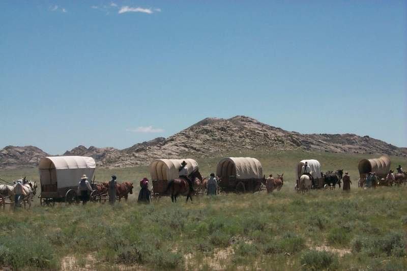 Tracing the Past: 6 Historic Trails In Wyoming 5 People stand beside covered wagons and horses in tall grass, with rugged mountains rising in the distance.