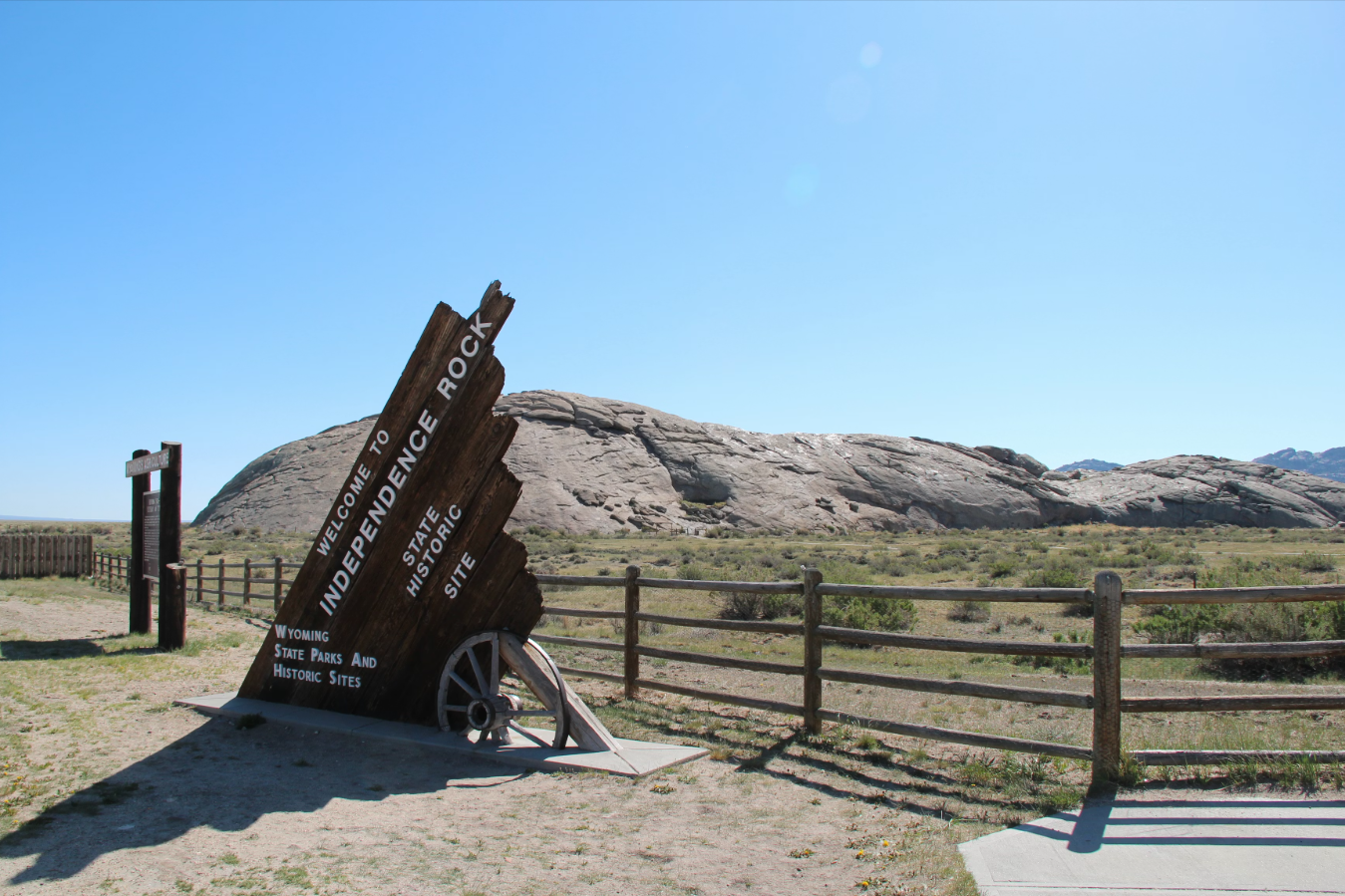 An entrance sign sits outside a wooden fence on the Wyoming prairie with rocks and mountains in the background. The sign reads: Welcome to Independence Rock, State Historic Site, Wyoming State Parks and Historic Sites.