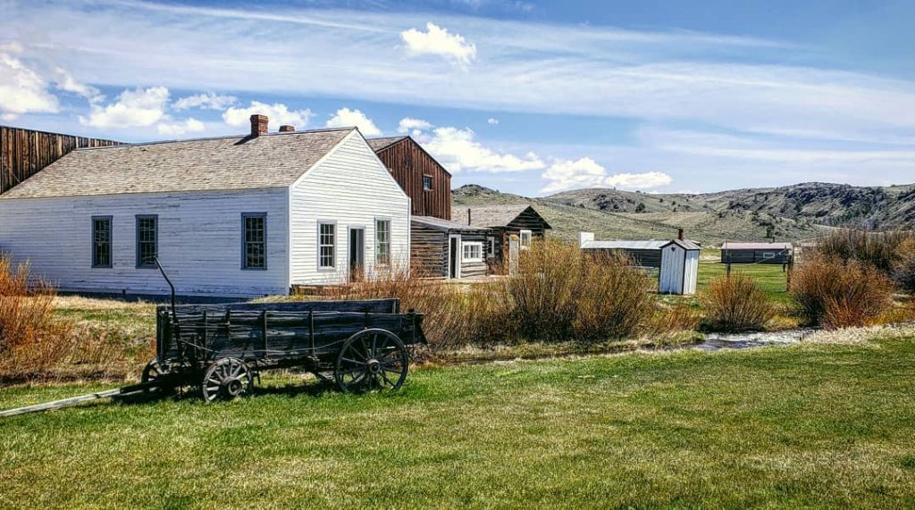A preserved pioneer-era town with wooden buildings and a vintage wagon in the grass under a bright blue sky in South Pass City, Wyoming.